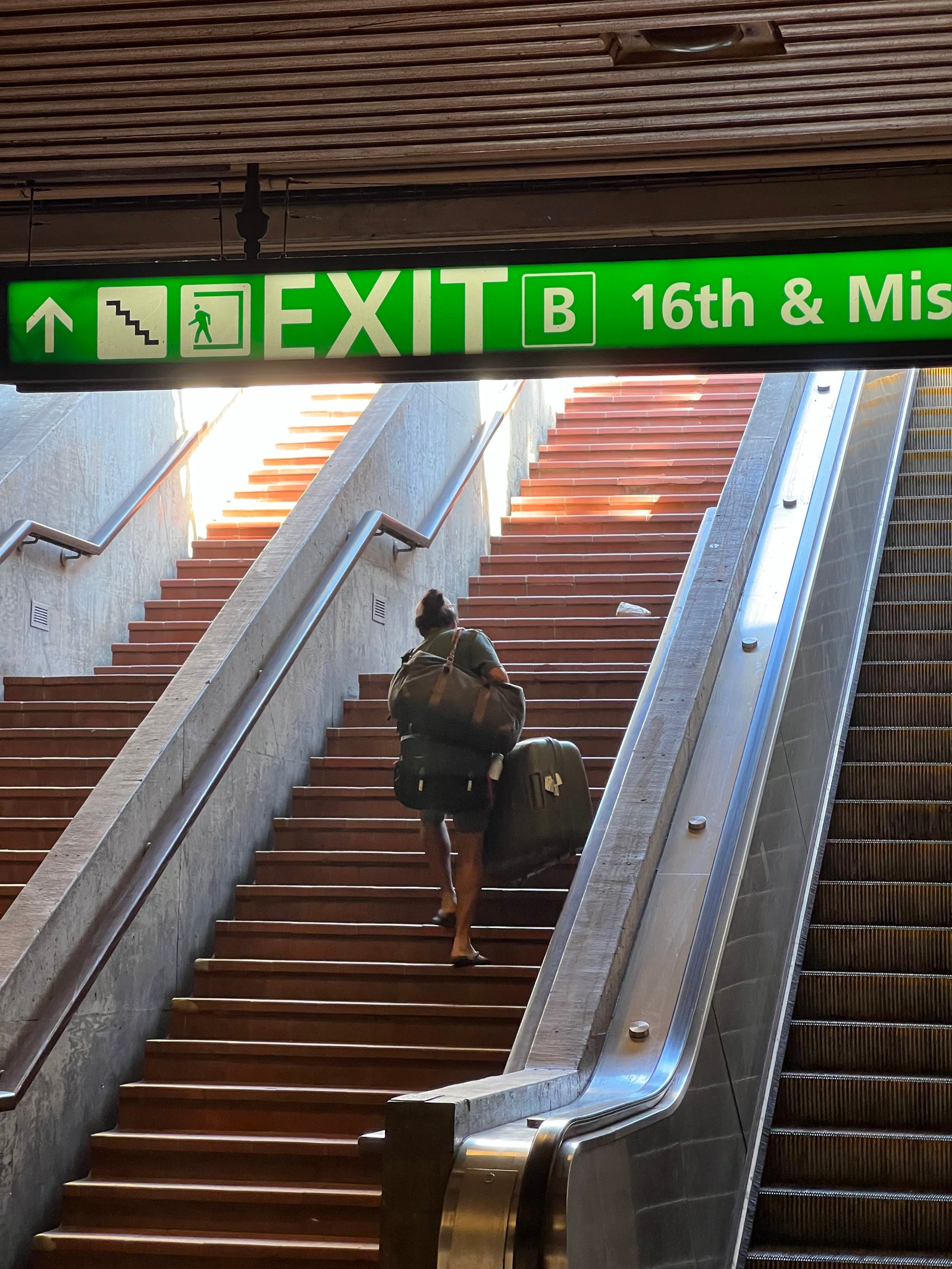 16th and Mission Exit B — a rider hauls heavy bags up the stairs beside the escalator